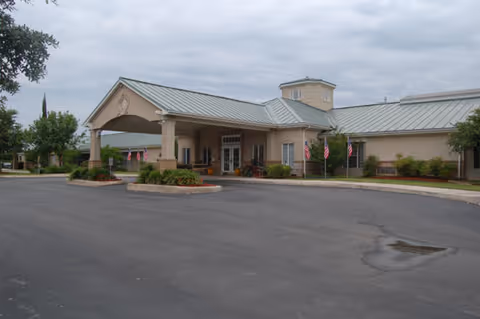 Entrance facade of a single-story senior living building with a covered porte-cochère, American flags, and a paved driveway under a cloudy sky.