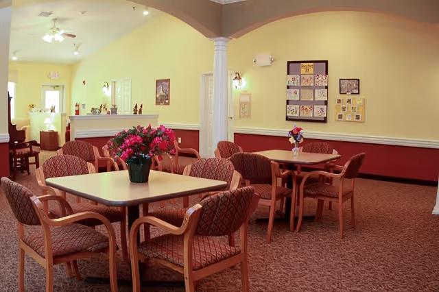 A cozy common area with several tables and cushioned chairs arranged around them. Each table has a vase with flowers. The walls are painted yellow and maroon, decorated with framed artwork and a bulletin board. There is a white column in the middle of the room and a ceiling fan above.