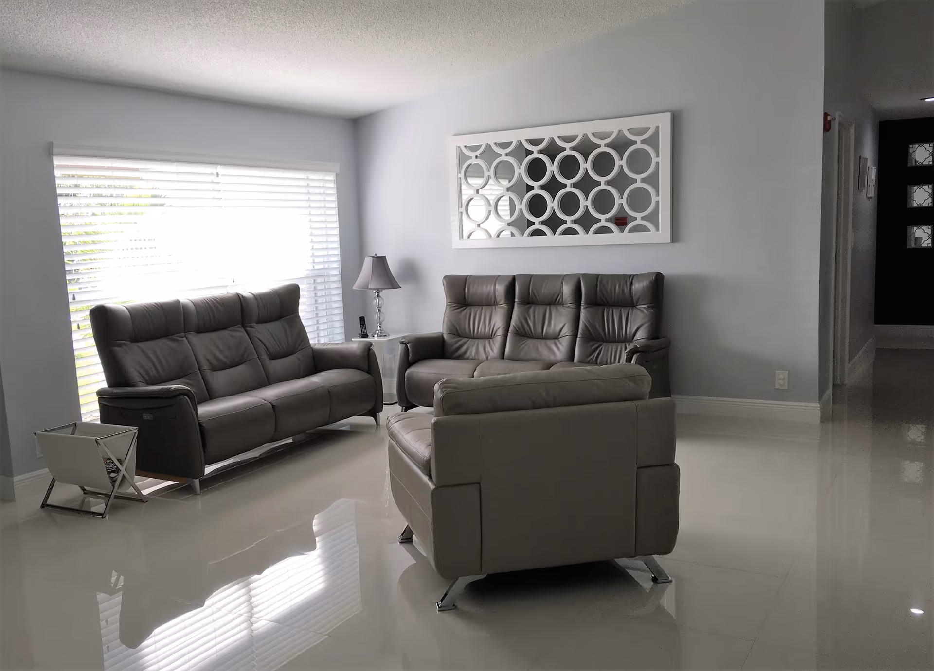A modern living room with two gray leather sofas and one matching armchair arranged on a shiny white tiled floor. A white side table with a lamp is placed between the sofas. A large window with white blinds lets in natural light, and a decorative mirror with circular patterns hangs on the light gray wall. A hallway with a dark door is visible in the background.