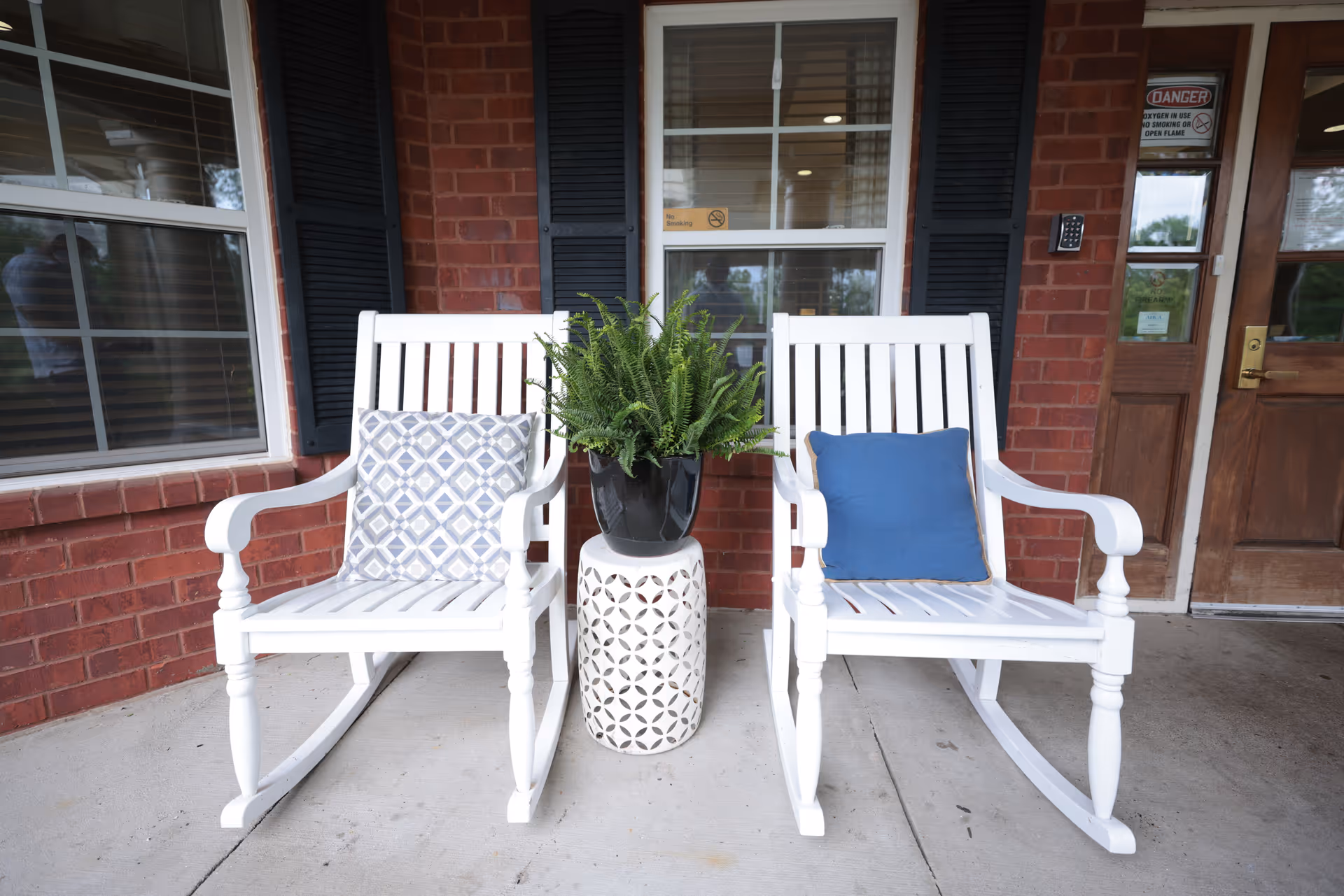 Two white rocking chairs with cushions flank a potted fern on a porch outside a brick building entrance.