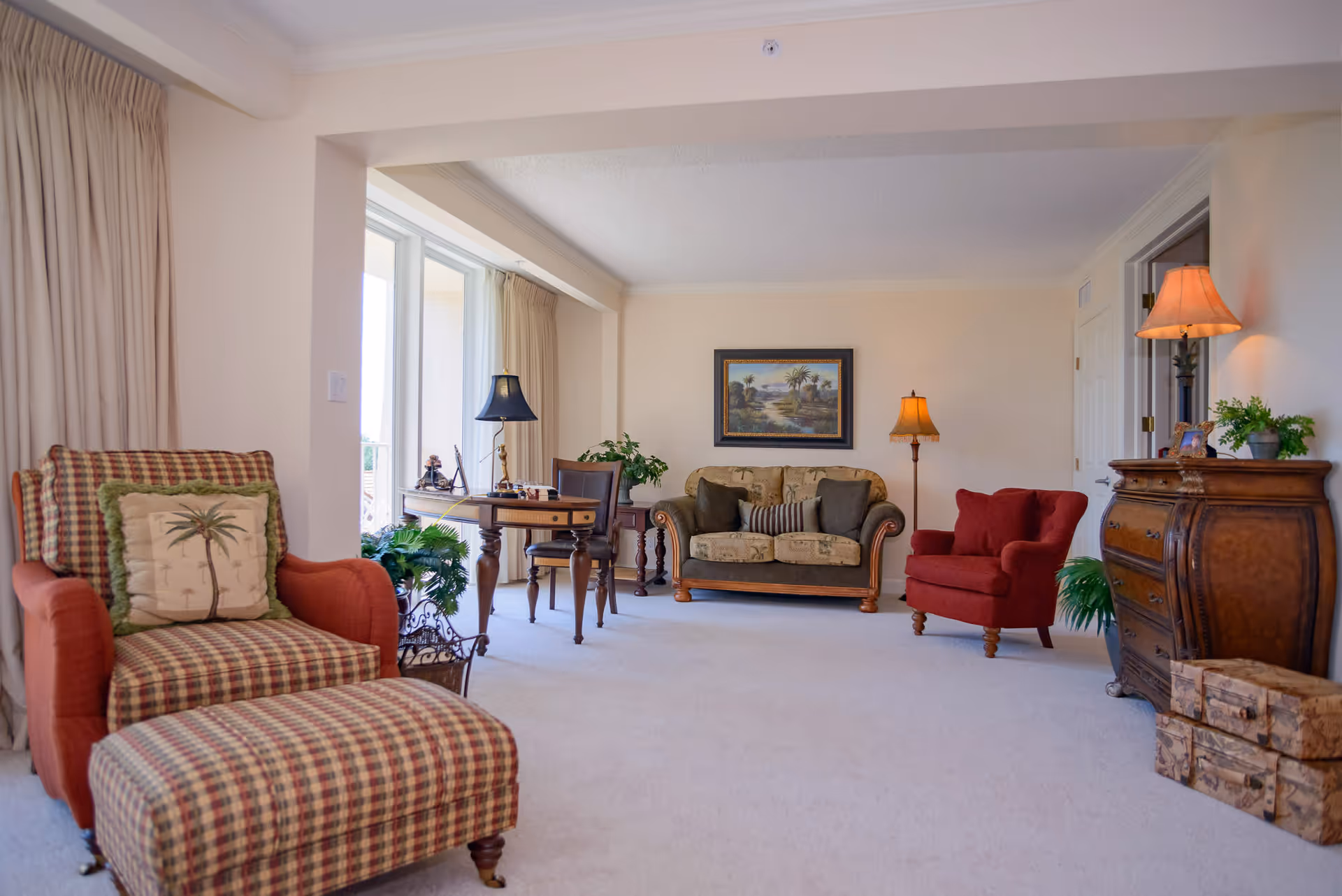 Sunlit traditional living room with patterned armchairs, a sofa, side tables, lamps, and potted plants.