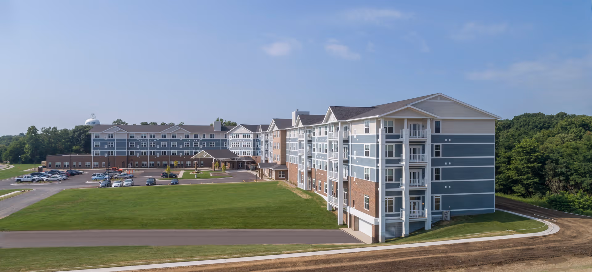 Exterior view of a large multi-story senior living facility building with blue and beige siding, white trim, and a covered entrance. The building is surrounded by a green lawn, parking lot with cars, and trees in the background under a partly cloudy sky.