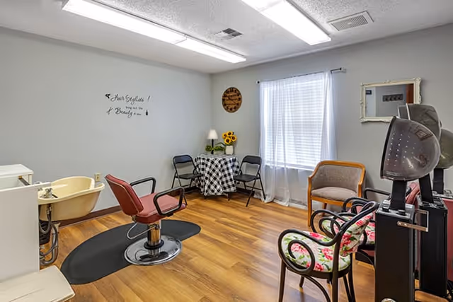 A bright room with wooden flooring featuring a salon chair in front of a small sink, a table with a checkered tablecloth and four chairs, a window with sheer white curtains, and a few decorative items including a wall clock, a lamp, and a vase with sunflowers.