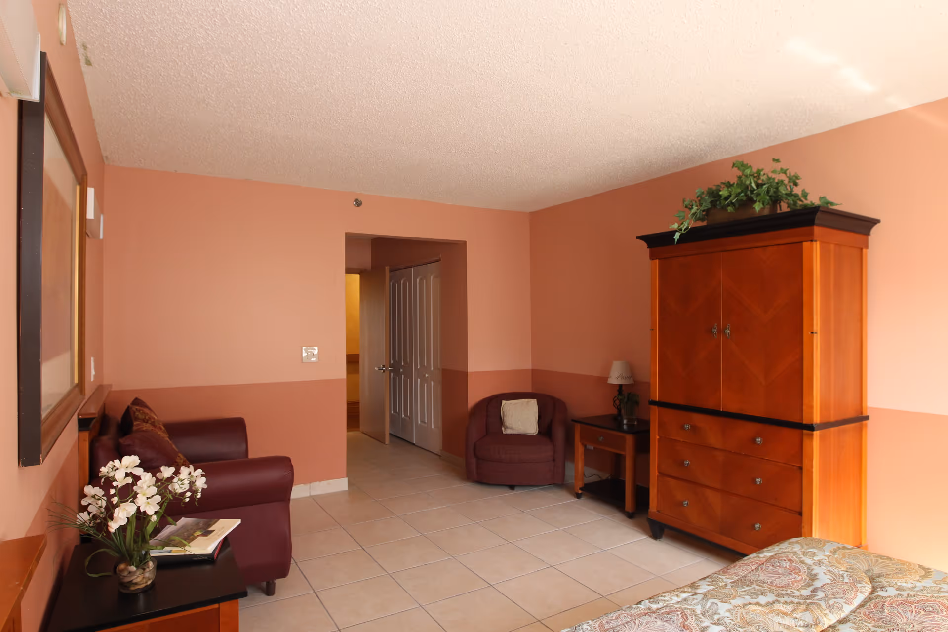 A cozy room with peach-colored walls and tiled floor featuring a maroon armchair, a wooden armoire with a plant on top, a side table with a lamp, a small sofa with cushions, a framed picture on the wall, and a bed partially visible with a patterned bedspread.