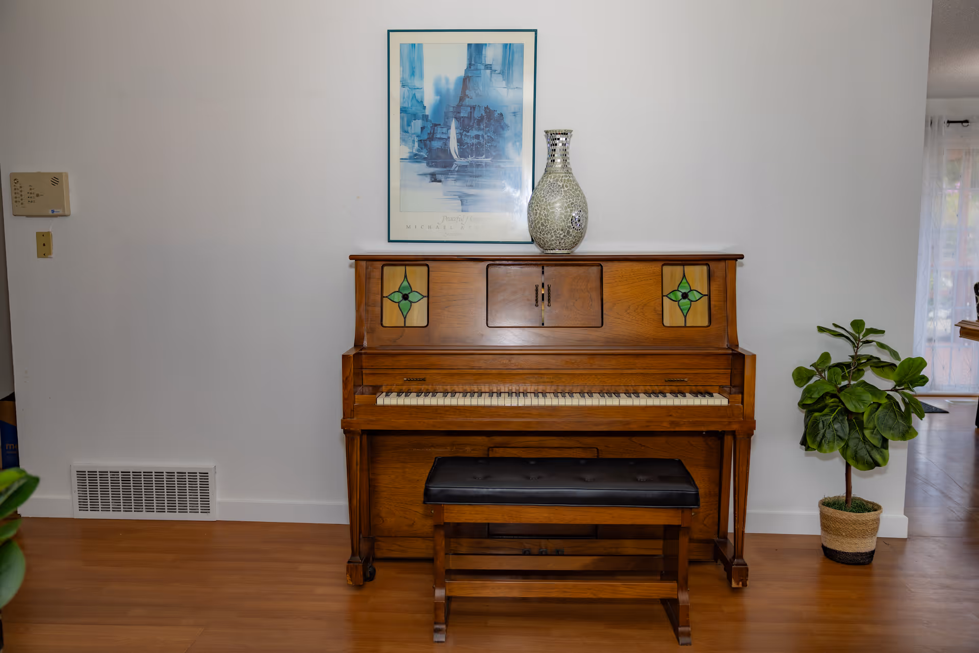 An upright wooden piano with stained glass panels on the front, a black cushioned bench in front of it, a decorative vase on top, and a framed blue-toned artwork hanging on the white wall above. To the right of the piano is a potted green plant, and the floor is wooden.