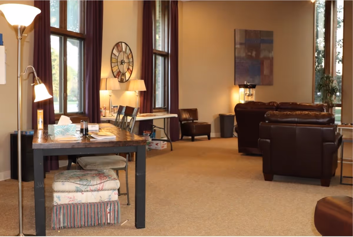 A cozy living room area with large windows covered by dark curtains, a wooden table with chairs, a floor lamp, and a wall clock. The room features brown leather armchairs and sofas, a side table with a lamp, and a piece of abstract wall art. The space is carpeted and well-lit with natural light coming through the windows.