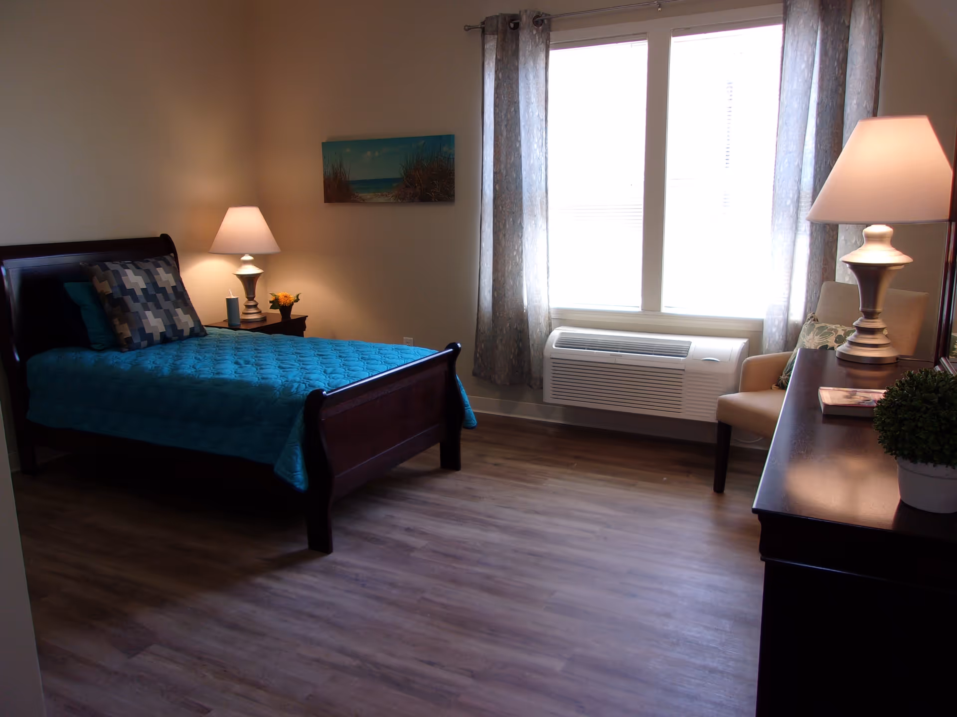A bedroom with a wooden bed frame and a teal quilted bedspread, two patterned pillows, a nightstand with a lamp and a small flower arrangement, a window with sheer curtains, an air conditioning unit below the window, a beige armchair with a decorative pillow, and a dark wooden dresser with a lamp, a book, and a small potted plant.