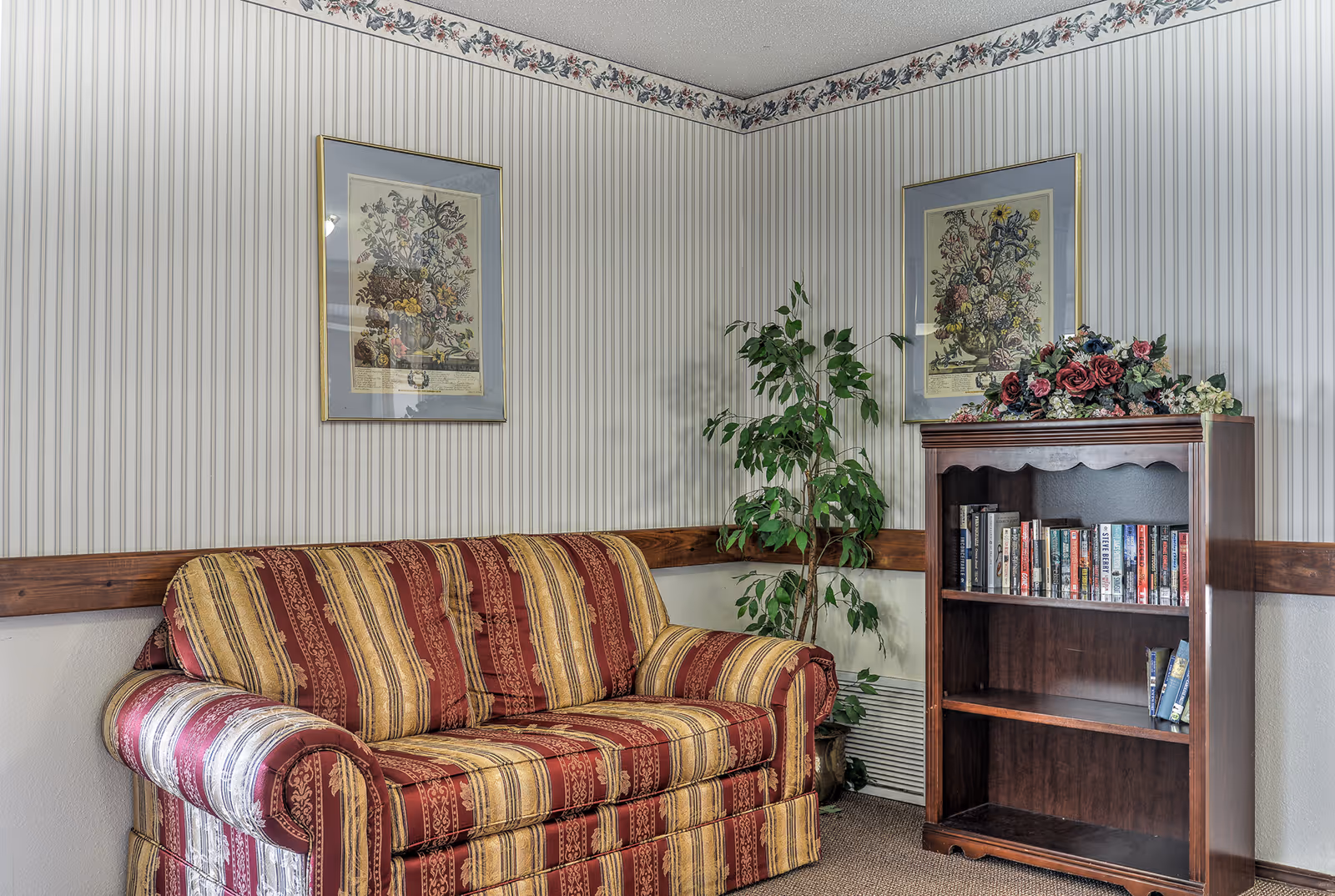 A cozy corner of a living room featuring a striped red and gold upholstered sofa, a wooden bookshelf with books and floral decorations on top, a potted green plant, and two framed floral prints hanging on striped wallpapered walls with a floral border near the ceiling.
