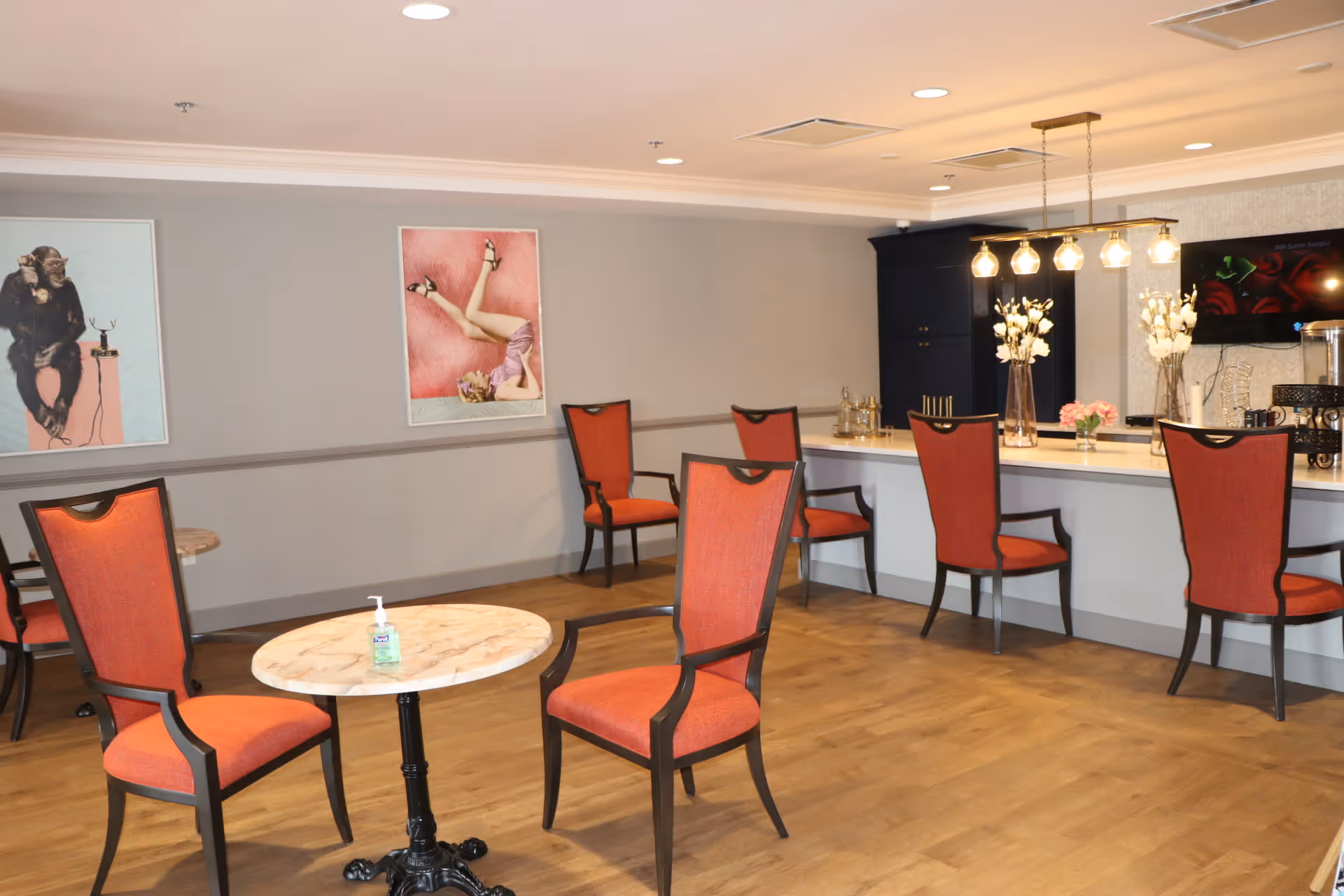 A well-lit interior room with wooden flooring featuring several red cushioned chairs around small round tables and a long counter with more chairs. The counter has decorative vases with white flowers and a coffee station. Two framed artworks hang on the gray wall, one depicting a chimpanzee with a telephone and the other a woman lying on a pink background. A hand sanitizer bottle is placed on one of the tables.