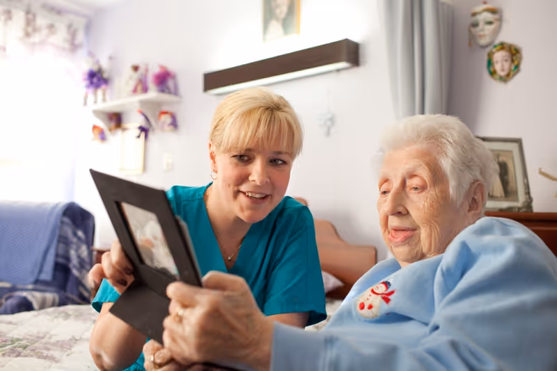 A female caregiver in teal scrubs smiling and showing a framed photo to an elderly woman wearing a light blue sweater with a snowman design, both sitting on a bed in a cozy room decorated with masks and framed pictures.