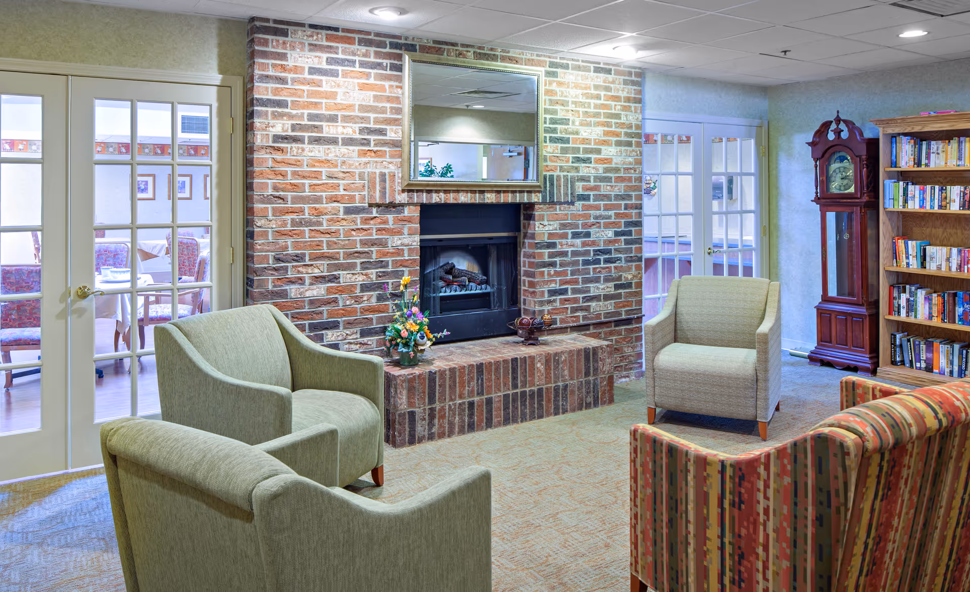 A cozy sitting area with a brick fireplace in the center, flanked by two beige armchairs and a patterned sofa. A wooden grandfather clock and a bookshelf filled with books are visible on the right side. Behind glass-paneled double doors on the left, a dining area with tables and chairs can be seen.