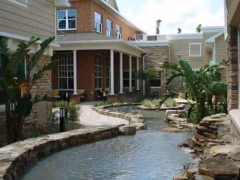 Outdoor courtyard with a stone-lined water feature between residential-style buildings and greenery.