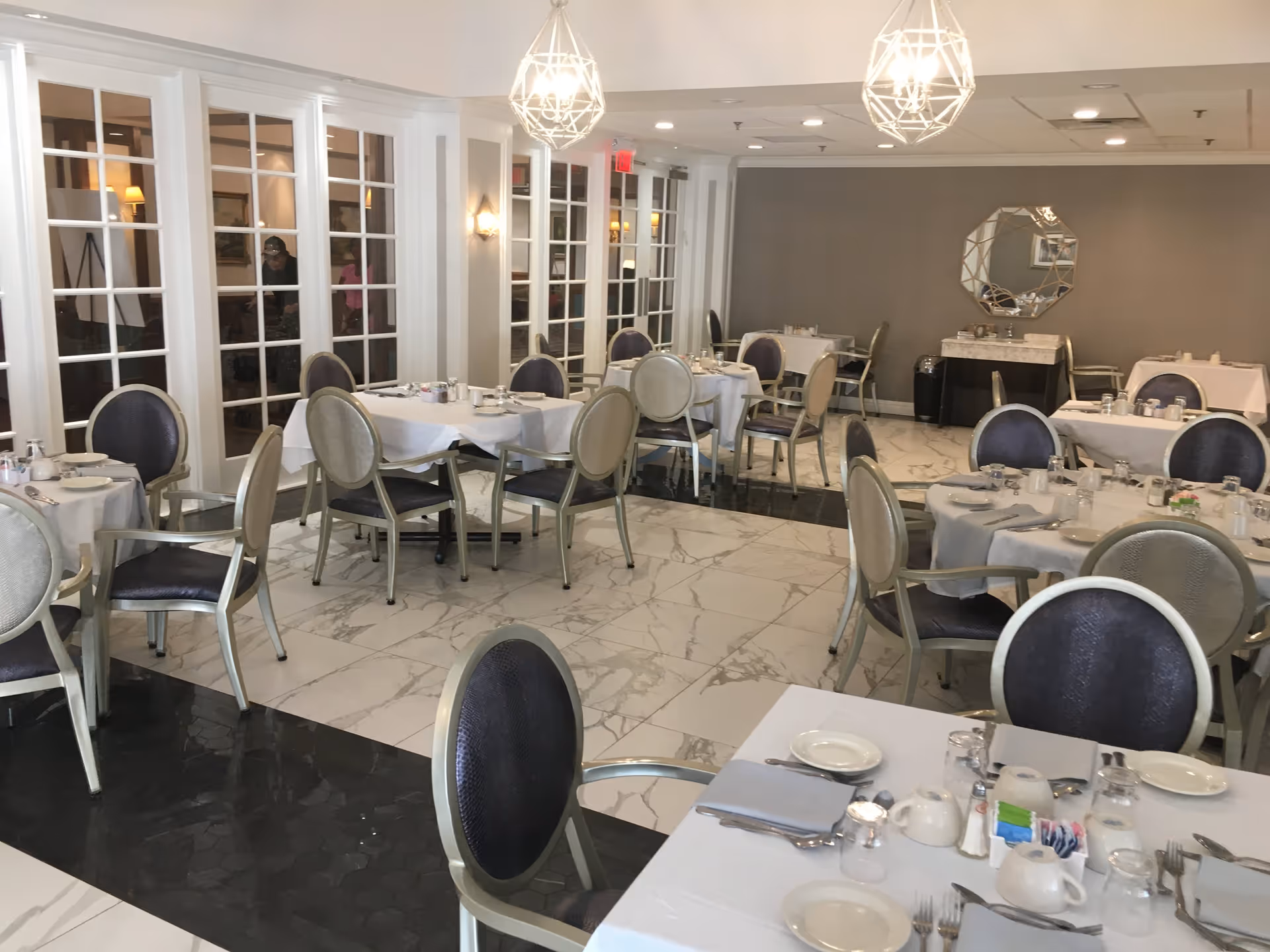 A bright and elegant dining room with multiple round and rectangular tables covered with white tablecloths. Each table is set with plates, cups, silverware, and condiments. The room features marble flooring with a mix of white and black tiles, cushioned chairs with dark seats and light frames, and decorative pendant lights hanging from the ceiling. A large mirror and a sideboard are visible against a gray wall in the background.