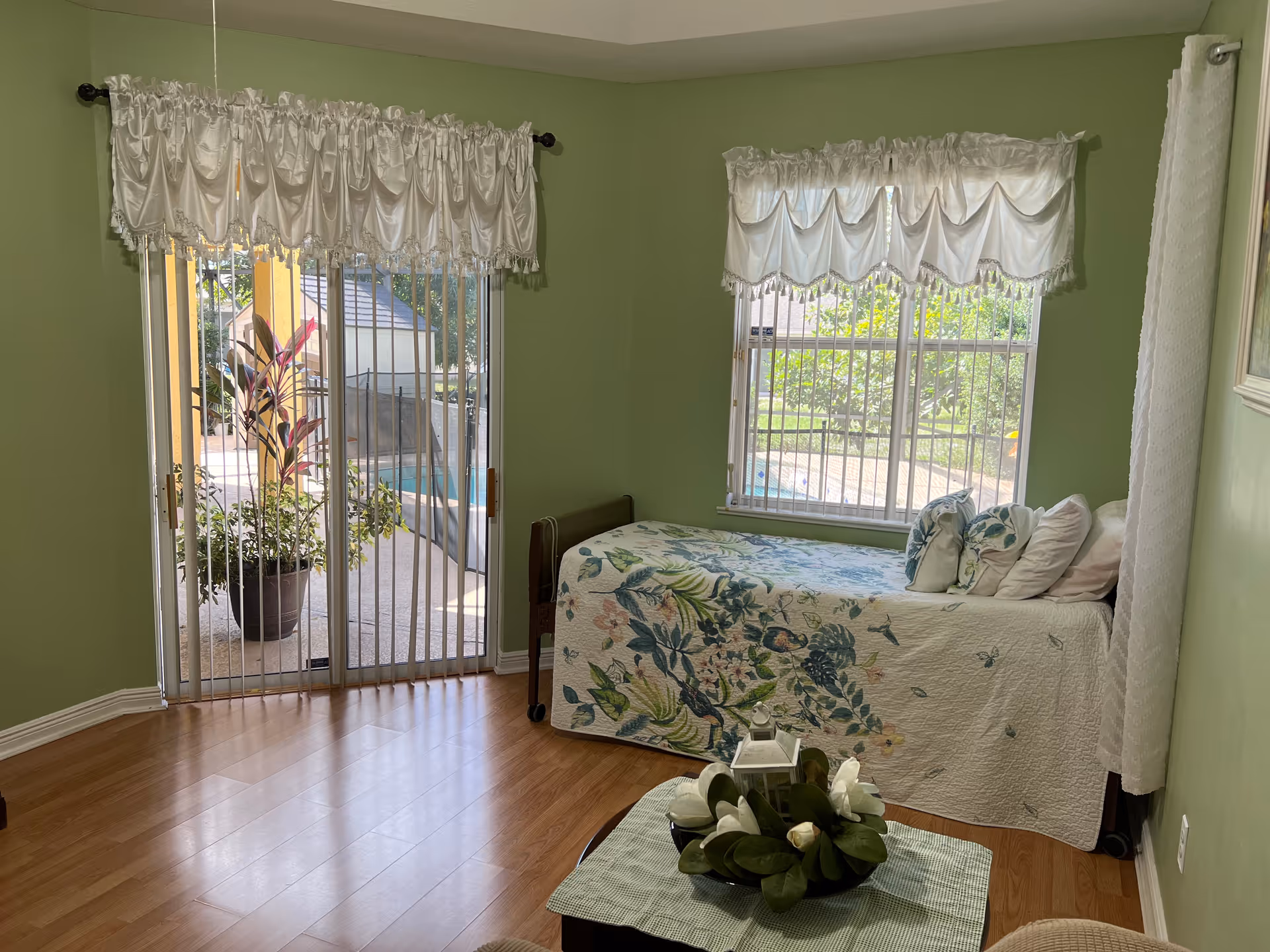A small bedroom with light green walls and wood flooring. There is a single bed with a floral bedspread and several pillows against the right wall. Two windows with white valance curtains let in natural light, and a sliding glass door with vertical blinds opens to an outdoor area with plants and a pool. A small table with a decorative centerpiece is in the foreground.