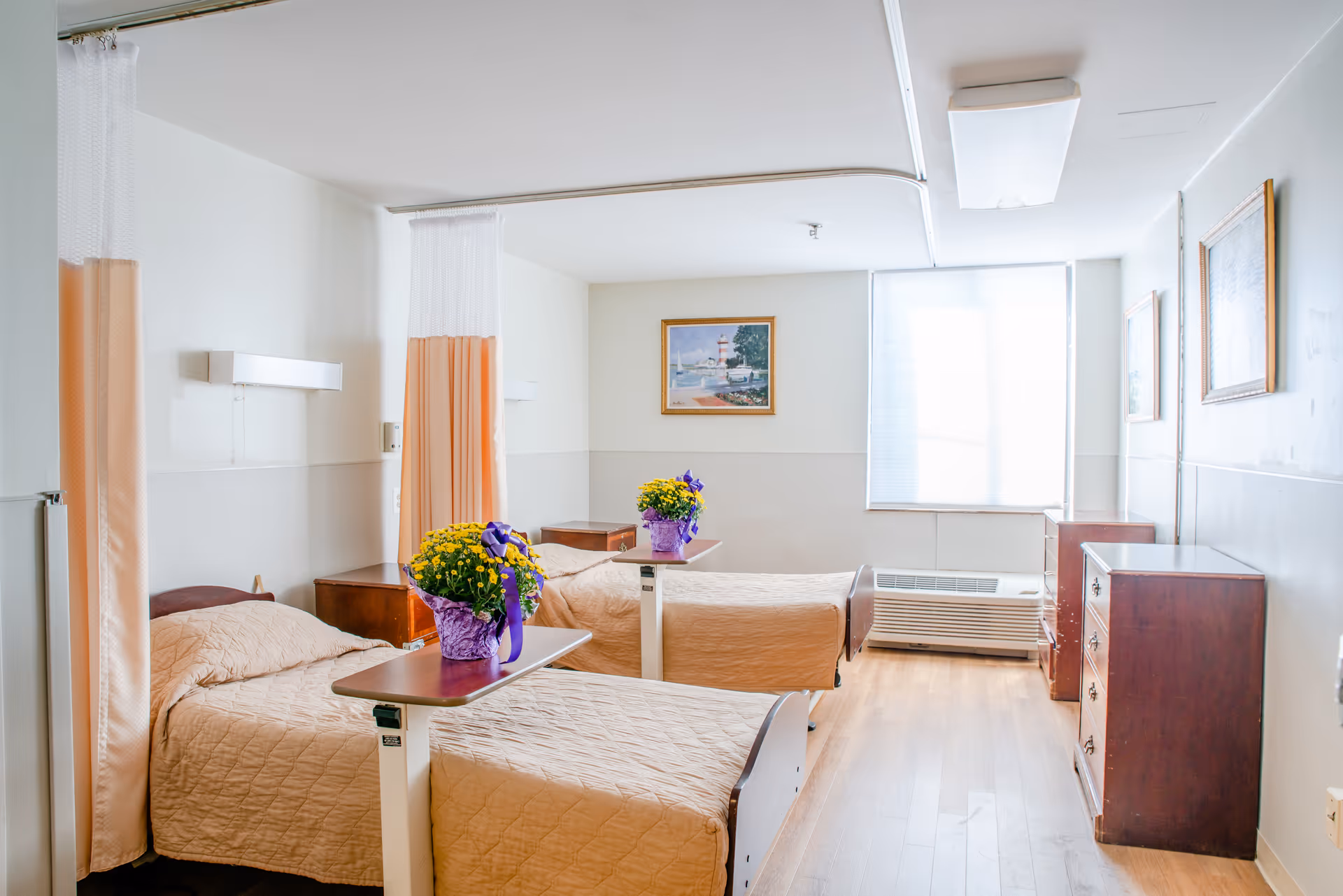 A bright room with two hospital-style beds separated by a privacy curtain. Each bed has a beige quilted blanket and a small overbed table with a potted yellow flower wrapped in purple paper. The room has light-colored walls, wooden furniture including dressers and nightstands, a large window with blinds, and framed artwork on the walls.