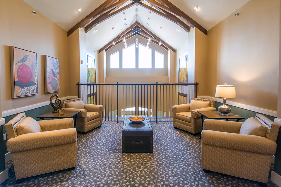 A cozy seating area with four beige armchairs arranged around a dark wooden coffee table with a decorative bowl. The space has a high vaulted ceiling with exposed wooden beams and large windows letting in natural light. Two framed artworks hang on the left wall, and a table lamp is on a side table to the right.