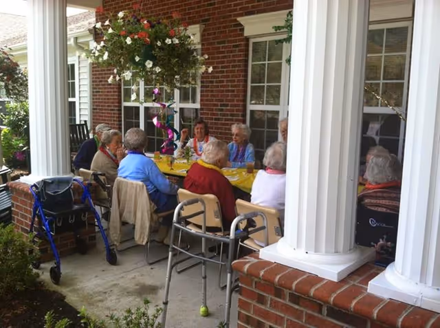 A group of elderly people sitting around a yellow table on a covered brick patio, engaged in conversation. The patio has white columns and hanging flower baskets, with walkers and wheelchairs nearby.