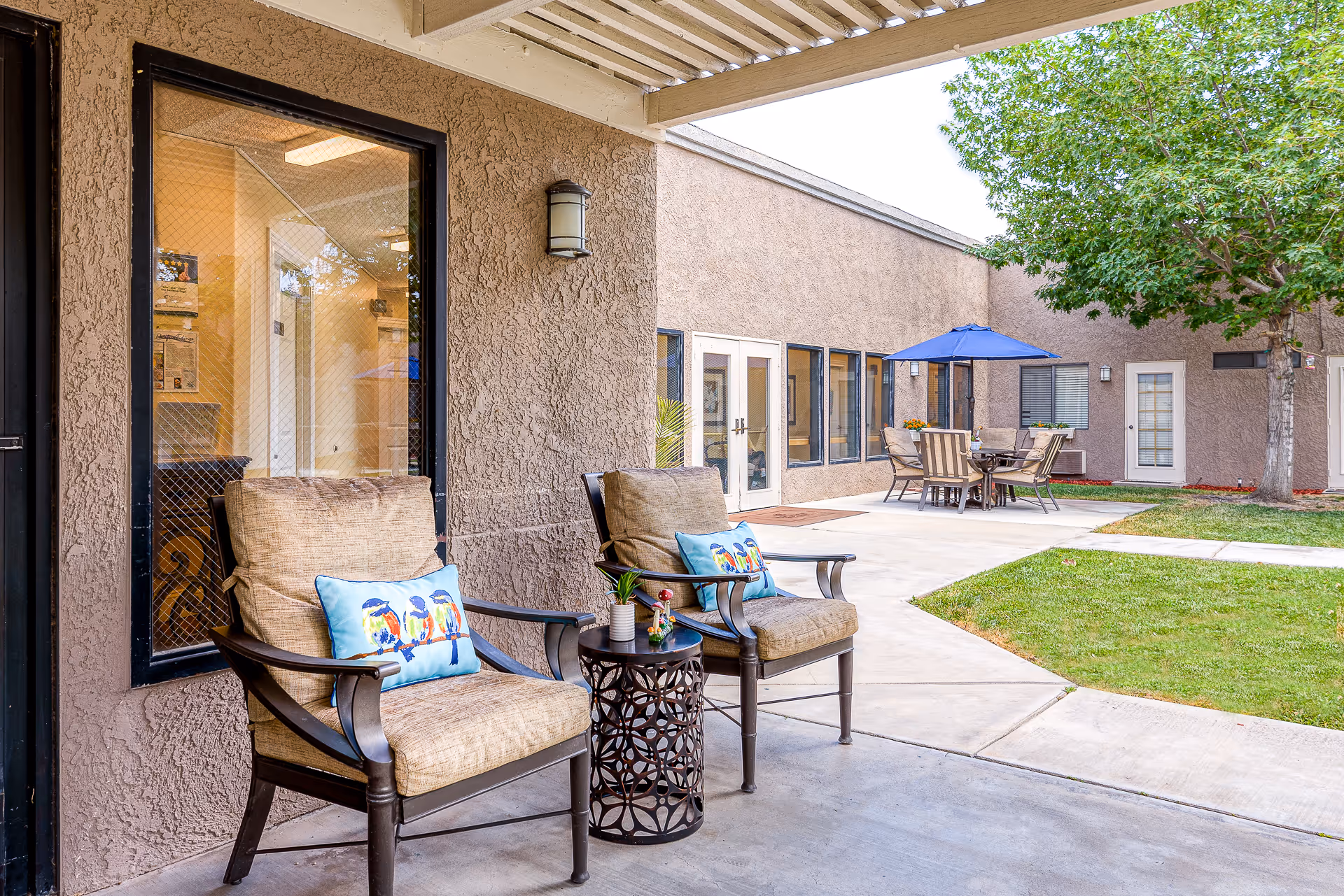 Outdoor patio area at Sierra Vista Independent & Assisted Living featuring two cushioned chairs with decorative bird pillows, a small round table with a plant, a concrete walkway, green lawn, a tree, and a dining table with chairs under a blue umbrella near the building.