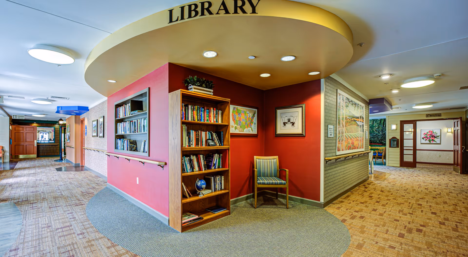 Interior view of a senior living facility hallway featuring a small library nook with a wooden bookshelf filled with books, a single striped chair, and framed artwork on red walls. The ceiling above the nook is curved and labeled 'LIBRARY'. The hallway is carpeted and has handrails along the walls, with additional artwork and doors visible further down the corridor.