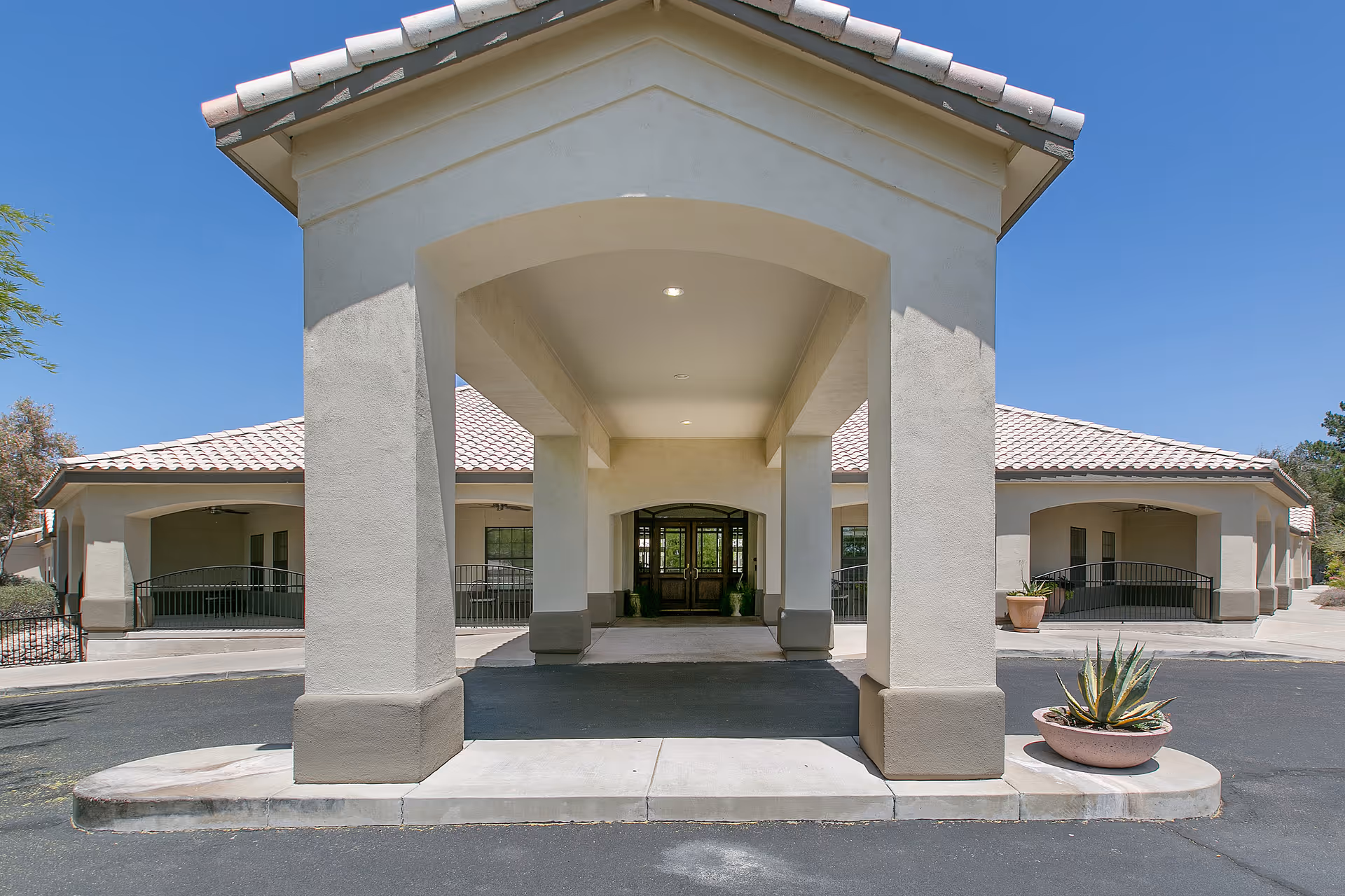 Front exterior view of a single-story building with a covered entrance supported by large columns, beige walls, and a tiled roof under a clear blue sky. There are potted plants near the entrance and a paved driveway in front.