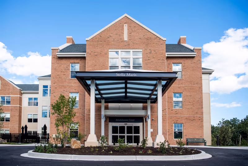 Front exterior view of the Stella Maris building, a multi-story brick structure with a covered entrance labeled 'Stella Maris' and 'Rehabilitation Center'. There are windows on each floor, some landscaping with small trees and bushes in front, and a clear blue sky with some clouds in the background.