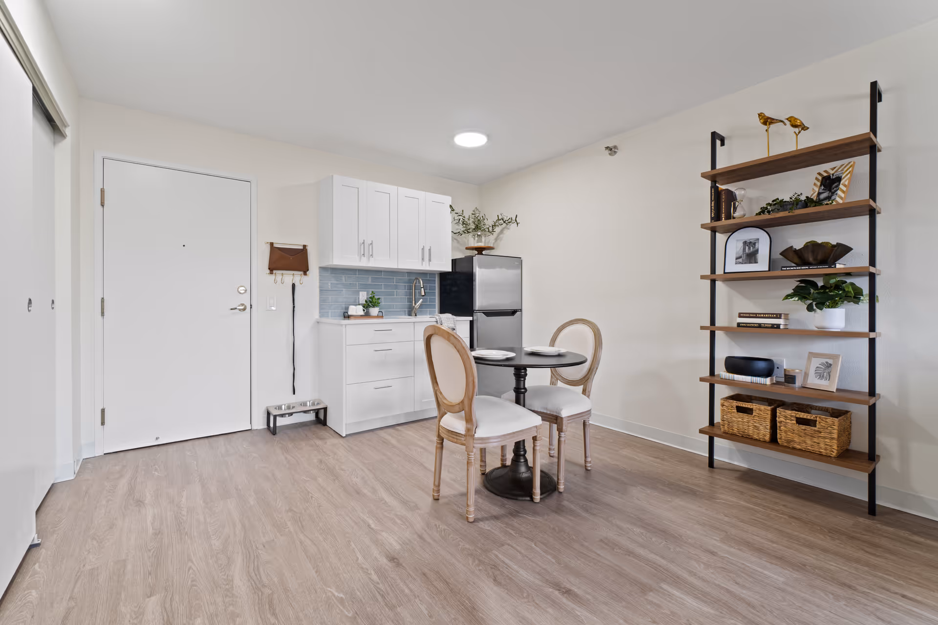 A small dining area and kitchenette in a senior living facility. The space features a round black table with two upholstered chairs, a white kitchenette with cabinets, a small refrigerator, and a blue tile backsplash. To the right, there is a tall shelving unit with decorative items, plants, and baskets. The floor is light wood, and the walls are painted white.
