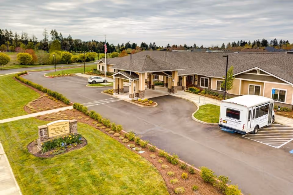 Aerial view of Arbor Oaks Terrace Memory Care Residence showing the building entrance with a covered drop-off area, a parking lot with a shuttle bus parked in a handicapped spot, landscaped green lawns, and a sign with the facility name near the road.