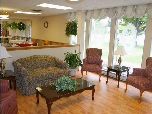 A cozy living room area with a patterned sofa, two upholstered armchairs, a glass-top coffee table with a green plant centerpiece, and two side tables with lamps. Large windows with decorative curtains provide a view of the outdoor greenery. The room has wooden flooring and hanging plants near the ceiling.
