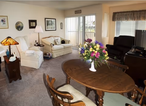 Combined living and dining area with a wooden dining table and floral centerpiece, upholstered sofa and chairs, recliner, TV, and sliding glass doors.