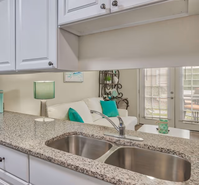 View of a kitchen sink with a granite countertop looking out into a living room area with a white couch, teal pillows, a teal lamp, and a glass door leading outside.