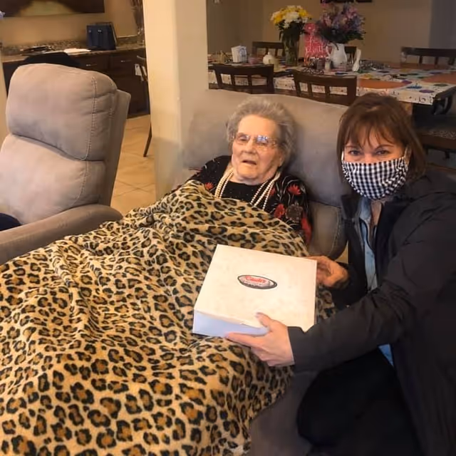 An elderly woman covered with a leopard print blanket sitting on a cushioned chair, accompanied by a younger woman wearing a checkered face mask and dark jacket, holding a white box. In the background, there is a dining table with chairs and a vase of flowers.
