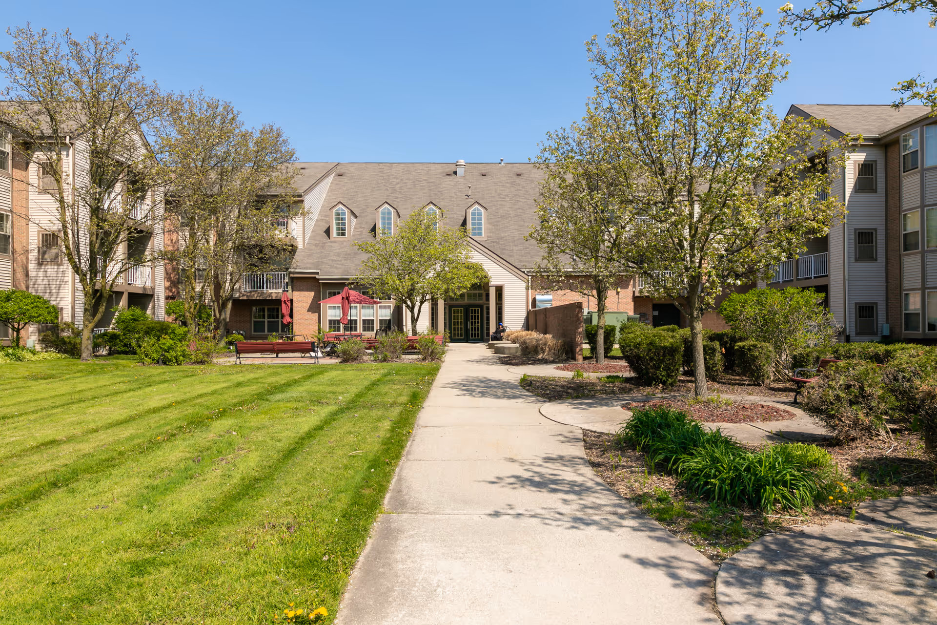 Front entrance and courtyard of a senior living building with a central walkway, green lawn, benches, trees, and multi-story residential wings.