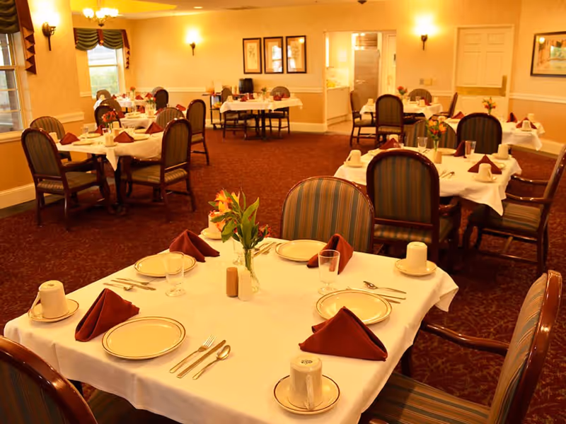 Warmly lit dining room with tables set with plates, folded napkins, glasses, and small floral centerpieces.