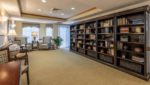 A cozy interior room with beige carpet and cream-colored walls featuring a large dark wood bookshelf filled with books and decorative items on the right side. On the left, there are several chairs, a globe on a stand, two table lamps, and windows with blinds and valances letting in natural light. The ceiling has recessed lighting and a wooden trim detail.