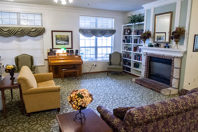 A cozy living room featuring two armchairs, a sofa, a fireplace with a stone surround, and a bookshelf filled with books. Natural light streams in through large windows with blinds, and a decorative flower arrangement sits on a coffee table.