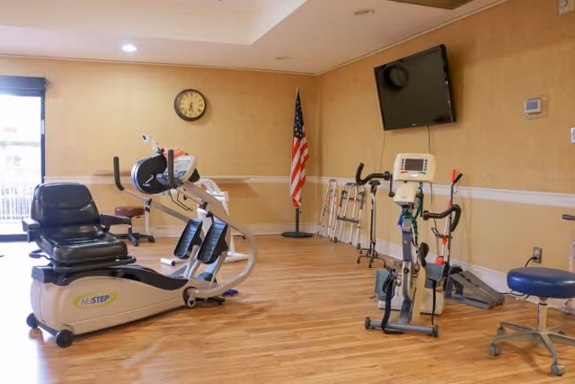 A rehabilitation room with exercise equipment including a NuStep recumbent cross trainer, a stationary bike, and other physical therapy tools. There are several walkers lined up against the wall, an American flag in the corner, a wall clock, and a mounted flat-screen TV. The room has wooden flooring and beige walls with a window letting in natural light.