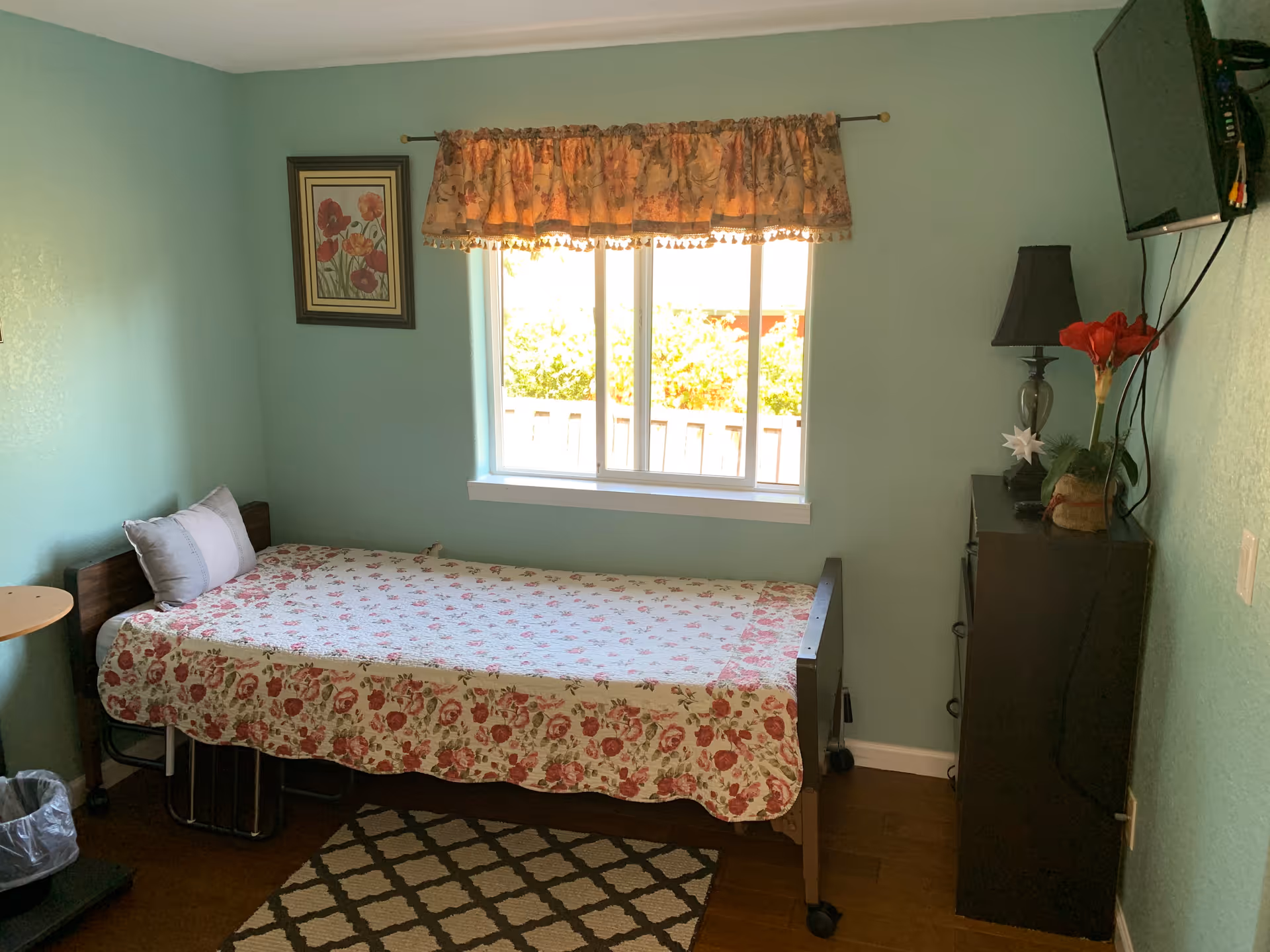 Single-occupancy bedroom with a floral-covered twin bed beneath a window, a wall-mounted TV, and a dresser with a lamp and flowers.