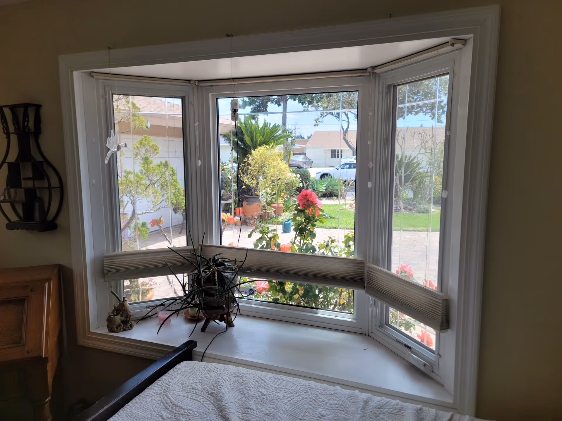 A bay window with white trim and partially lowered blinds looking out onto a garden with various plants and flowers. Inside, there is a windowsill with potted plants and a bed with a white quilted bedspread partially visible in the foreground. The room has light-colored walls and a wooden headboard is partially visible on the left side.