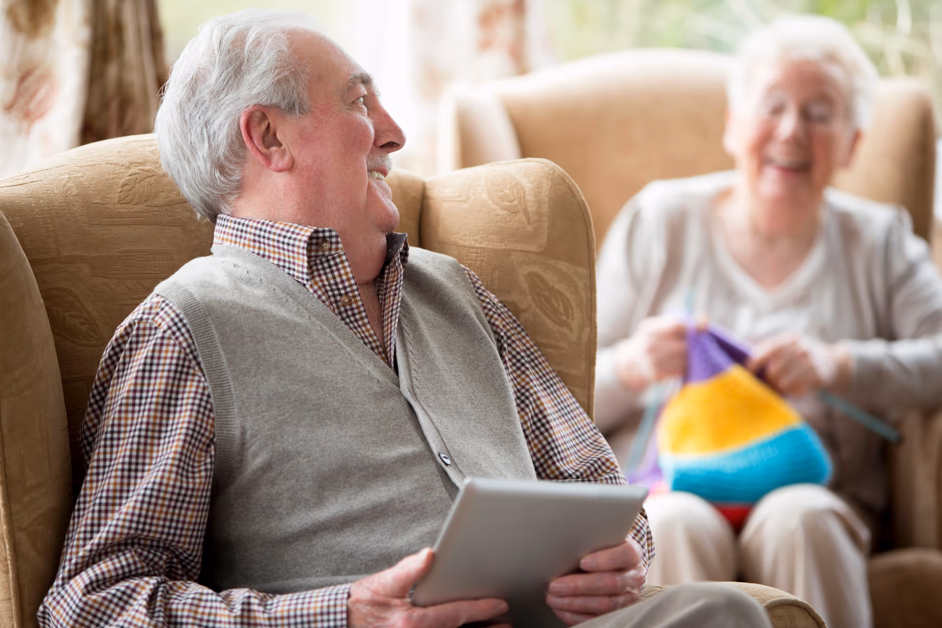 An elderly man sitting in a beige armchair holding a tablet, smiling and looking towards an elderly woman who is knitting with colorful yarn in the background, both in a cozy living room setting.