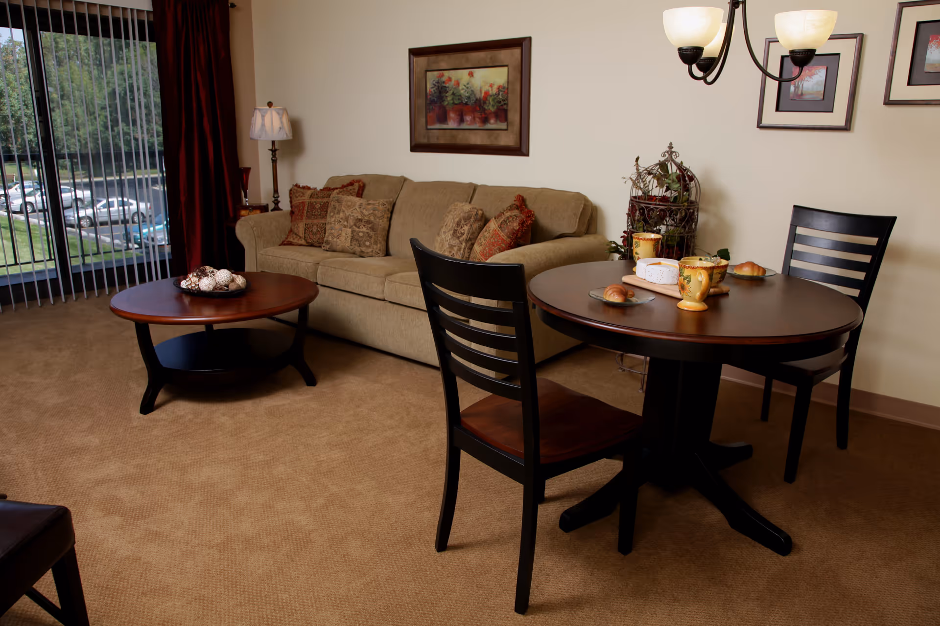 A cozy living room area with a beige sofa adorned with patterned cushions, a round wooden coffee table with decorative balls, and a small round dining table set with two chairs, plates with croissants, cups, and a cheese board. The room has beige carpet, framed artwork on the walls, a floor lamp, and vertical blinds covering a large window showing a parking lot outside.