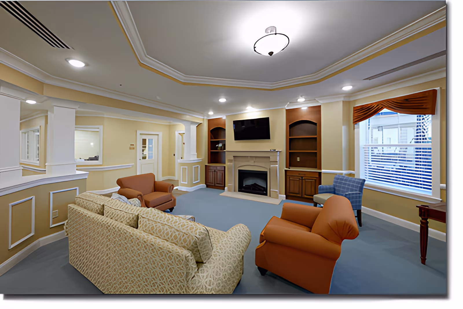 Bright senior living lounge with sofas and armchairs arranged around a fireplace and wall-mounted TV.