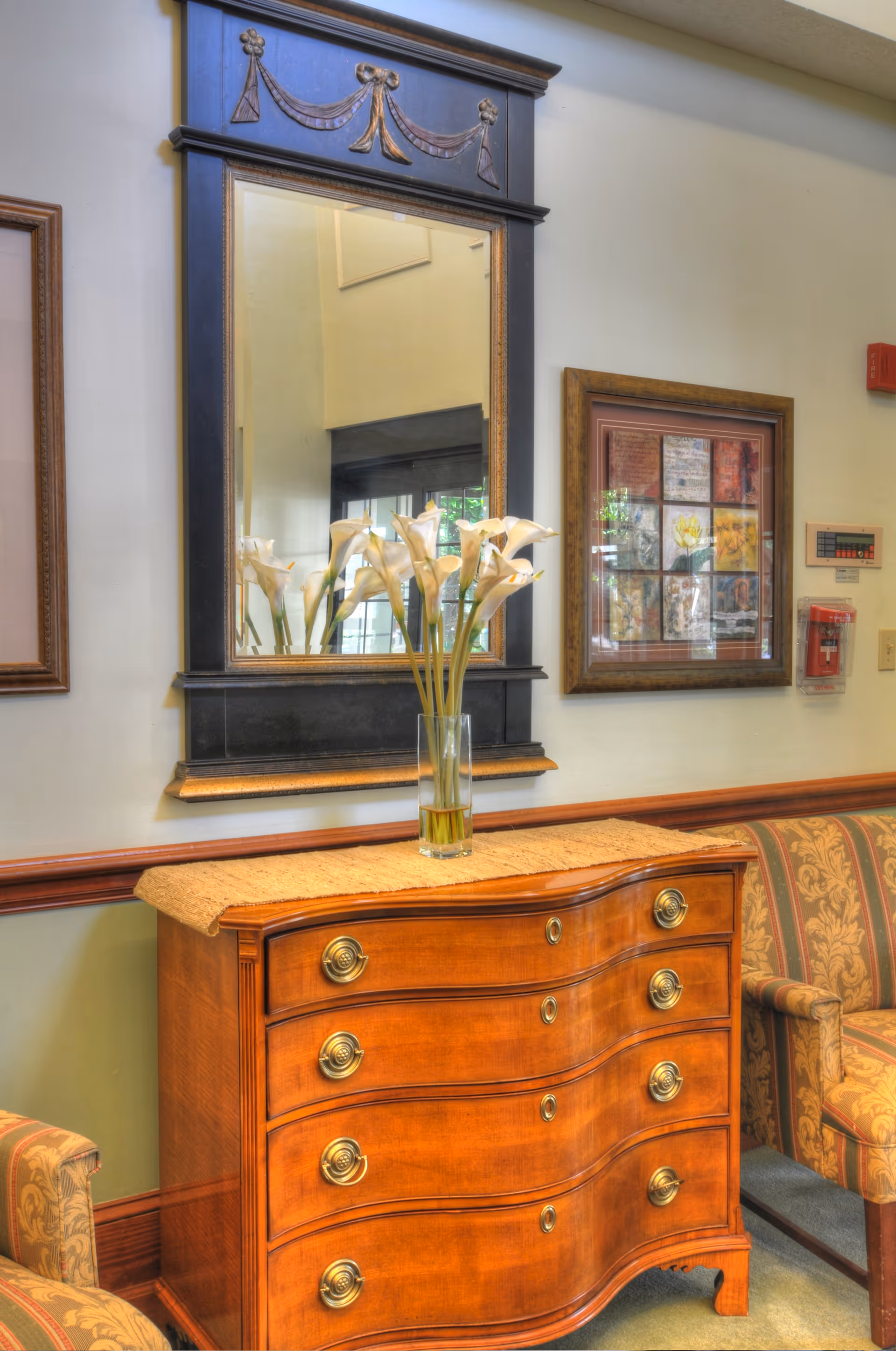 A wooden chest of drawers with curved front and brass handles, topped with a beige runner and a glass vase holding white calla lilies. Above the chest is a large decorative mirror with a black and gold frame. To the right, there is a framed collage artwork on the wall and a patterned upholstered armchair partially visible. The setting appears to be a cozy interior space with soft lighting.