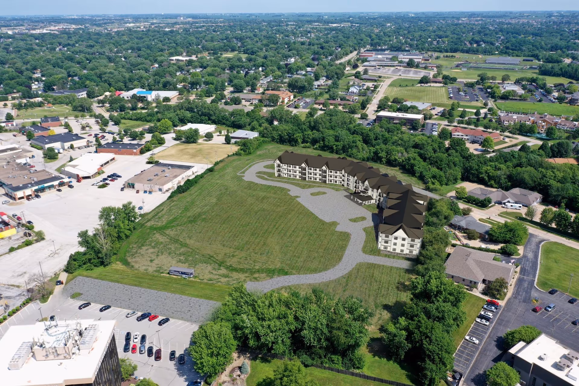 Aerial view of a suburban area showing a large grassy plot with a digitally rendered building complex labeled The Suites of Bettendorf. Surrounding the plot are trees, parking lots, roads, and various commercial and residential buildings extending into the distance under a clear sky.