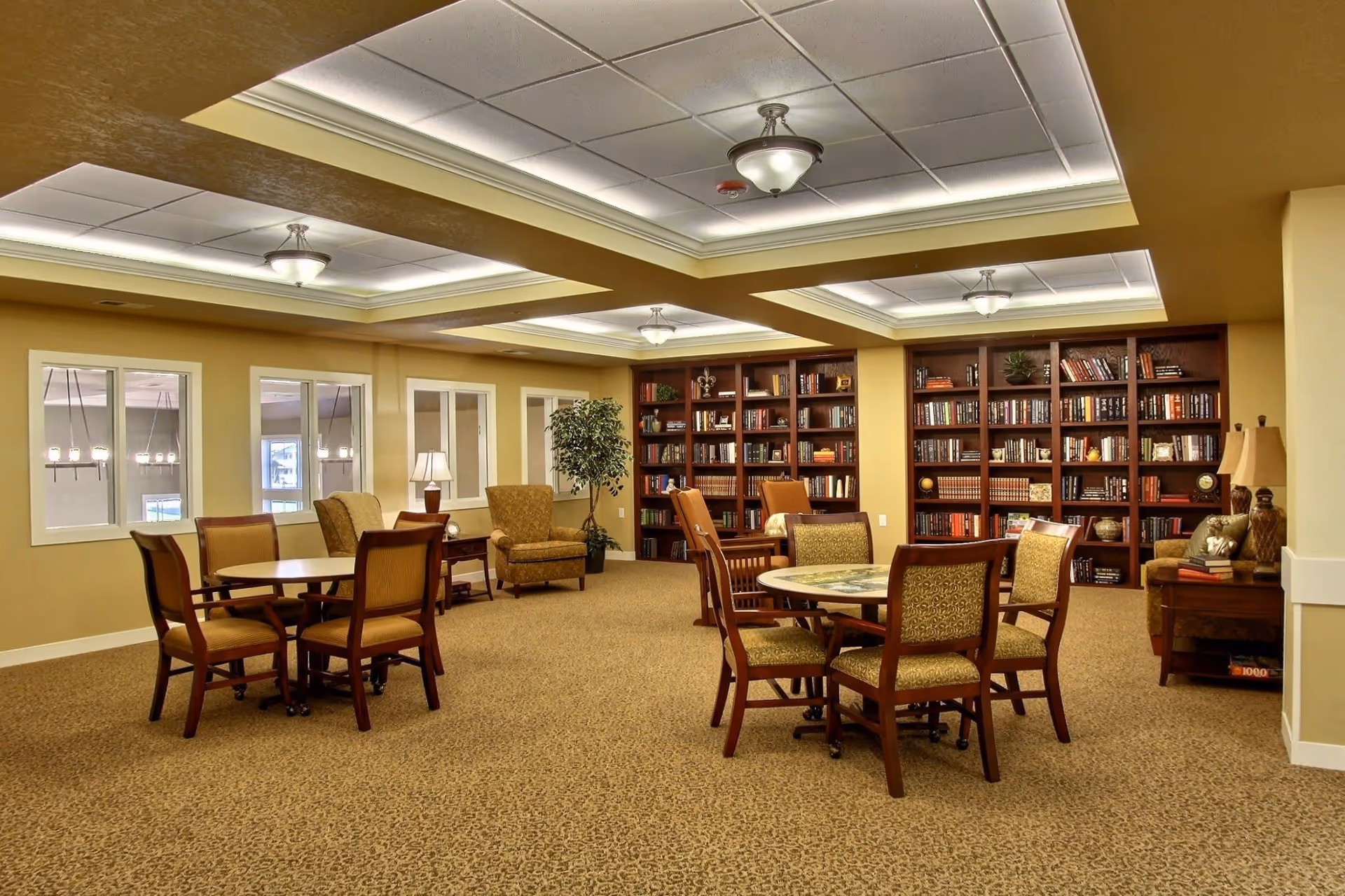 Well-lit senior living common room with round tables and chairs, armchairs, and a large bookshelf along the back wall.