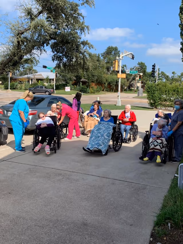 A group of elderly people sitting in wheelchairs outside on a sunny day, some covered with blankets. Several caregivers in colorful scrubs are attending to them. The setting appears to be a parking lot or driveway near a street intersection with trees and cars in the background.