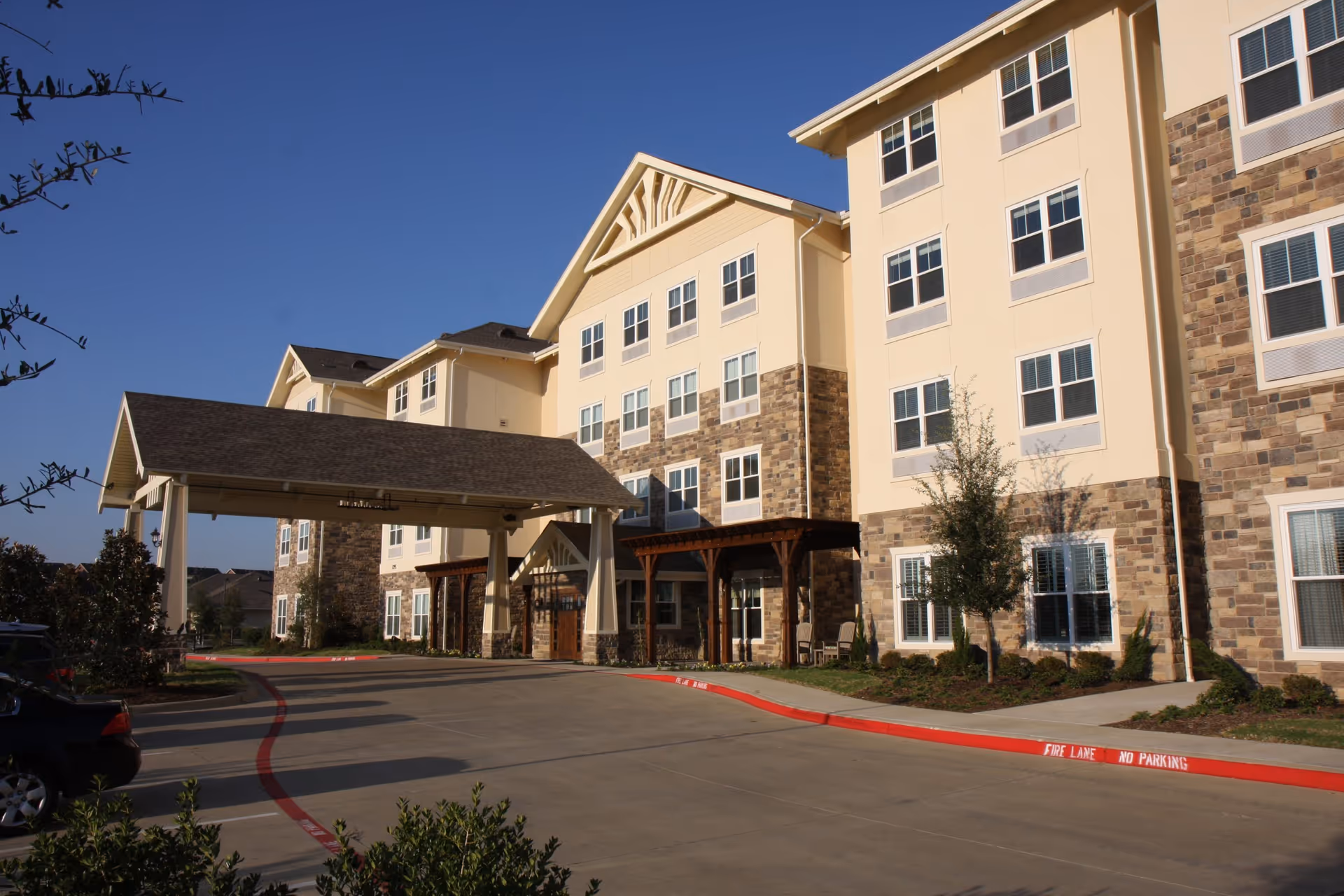 Front entrance of a multi-story senior living building with a covered porte-cochere, stone and beige siding, and a clear blue sky.