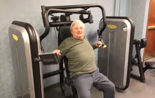 An elderly man using a seated chest press exercise machine in a fitness room with gray flooring and light-colored walls.
