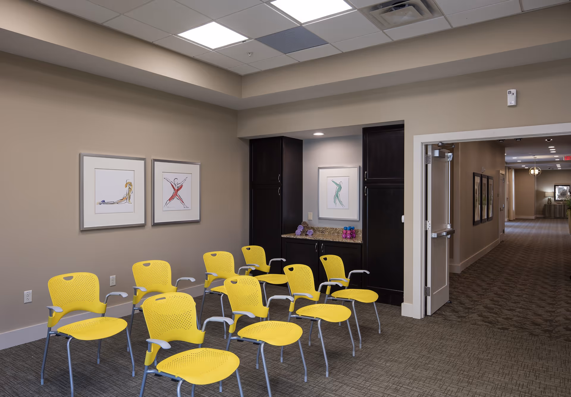 A room with eight yellow chairs arranged in rows facing forward. The walls are beige with three framed abstract art pieces. There is a dark wood cabinet with a granite countertop holding small purple and pink dumbbells. An open doorway leads to a hallway with more framed pictures and lighting fixtures.