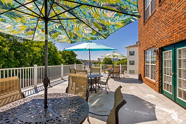 Outdoor terrace with metal tables and chairs under colorful umbrellas beside a brick building overlooking trees.