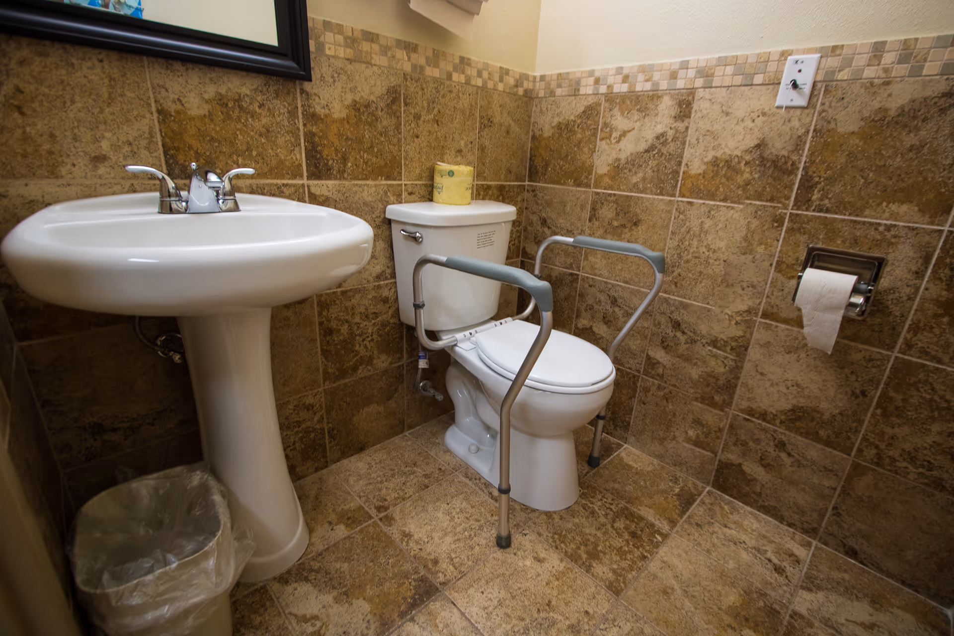 A bathroom with brown tiled walls and floor, featuring a white pedestal sink, a white toilet with safety rails on both sides, a toilet paper holder with a roll of toilet paper, and a small trash can with a plastic liner.